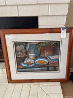 Photo showing the entire framed wall art standing on a white brick hearth. The frame is wood with a cream mat surrounding the print. The artwork depicts a still life with various food items, a glass bottle, plants, and stacked books.