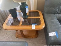 Top angled view of the side table showing glass panel inserts and wood frame, placed between couch cushions with a lamp on top.