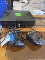 Black original Xbox console with two black wired controllers on wooden surface, cables visible.