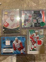 Four hockey cards featuring Detroit Red Wings players shown on table. Cards show players Gustav Lindstrom, Pavel Datsyuk, Steve Yzerman, and Ryan Kuffner with embedded jersey swatches and card numbering.