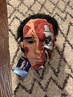 Front view of Terminator 2 mask showing half human face and robotic endoskeleton side with damage detailing and attached tag.