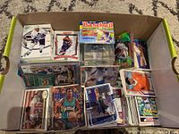 Top-down view of a box containing various sports cards including hockey, basketball, football, baseball, slabbed card, and unopened sticker pack