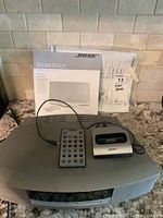 Photo showing main speaker unit, remote control, docking station, and user manual with original packaging in background.