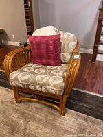 Rattan chair with beige and white leaf-patterned cushions and red throw pillow, on a brown and beige area rug, hardwood floor.