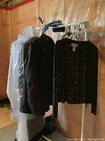 View of rack with multiple clothing items, including camouflage coat, dark coat, white garment in cover, black and silver patterned sweater top