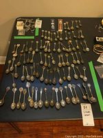 Overview of various collector spoons laid out in rows on a black table, showing their design variety and quantity.