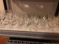 Photo showing a collection of various clear glass wine glasses, sherry glasses, and smaller glass items arranged in rows on a cart.