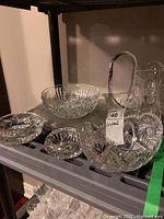 Crystal platter, bowls, ashtrays, pitcher, and basket arranged on a gray shelving unit, showing detailed cut glass patterns and shapes.