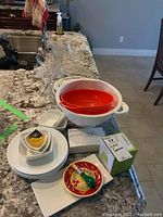 Photo showing plastic colanders in white and red, plastic bowls, small condiment dishes, a decorative plate with a fruit design, and boxes for spiralizer and digital thermometer.