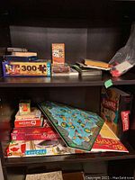 Shelf containing several boxed board games (Scrabble, WordQ, Triazzle, Toronto in a Box), card games (cribbage), books, and a puzzle set.
