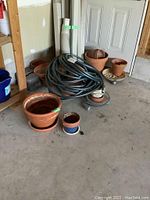 Coiled black garden hose with assorted terracotta and plastic planters on concrete floor.