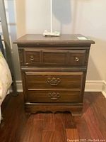 Front view of one dark wood nightstand showing two drawers with decorative hardware and worn surface.