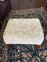 Full top and side view of beige floral patterned upholstered ottoman on a floral rug.
