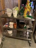 Photo of metal cart with various ceramic and pottery planters, some gardening supplies including soil and fertilizer bags on top shelf and terraces on bottom shelf.