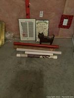 Four posters including a large framed one, three poster tubes, metal cat decor, painted stone welcome sign, and a wall plaque on concrete floor.