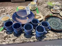 Overview photo showing large bowl, six mugs in front, extra mug, and signed condiment dish in background on countertop.