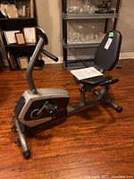 Side view of recumbent bike showing seat, pedals, and manual on seat; bike in a room with shelving and wood floor