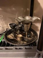 Large round silver plated tray, two goblets, scalloped dish, salad serving utensils, and tea strainer arranged on shelf