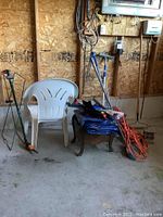 Overall view of plastic chair, rake, pruning shears, tarp, extension cord, small wood stool, and shovel leaning against wall
