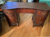 Front view of an antique mahogany vanity desk with a curved front. Shows multiple drawers on both sides and a center drawer. Some drawer handles are missing, and the wood surface displays wear and age.