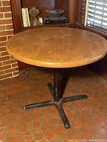 Side angle showing the round wooden tabletop and cast iron base with four legs on tiled floor near a window and bookshelf.