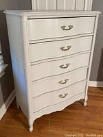 Front angled view of the painted chest showing drawer fronts and ornate brass handles, standing on hardwood floor against wall.