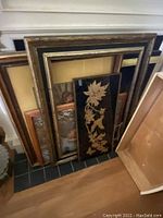 Multiple vintage picture frames stacked against a wall, showcasing variety in size and color including a distinctive black frame with gold leaf botanical design.