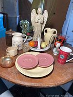 Overview of lot showing ceramic plates, angel figurine, various McCoy and Fiesta Ware pottery, basket with faux fruit, Starbucks mug and other items on wooden table.