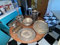 Overview of the lot displaying multiple vintage silver plated items on a wooden round table including trays, pitchers, and small kitchenware.