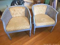 Front view of the two vintage painted wooden chairs with cane sides and beige fabric cushions.