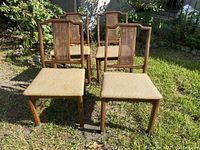 Front view of 4 mid-century dining chairs showing wooden frames, cane backrests, and beige fabric seats placed on grass outdoors.
