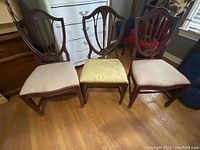 Three vintage mahogany chairs including two matching shield-back chairs with beige seats and one different chair with rounded back and yellow upholstered seat.