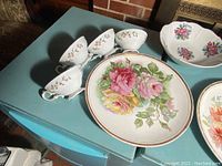 Photo showing five small porcelain cups with floral patterns and a large round plate with pink, yellow, and green roses painted in center with gold rimmed edge