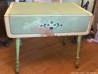 Image showing side view of vintage painted kitchen table with cream and green paint and decorative motif on apron. Some paint chipping is visible.