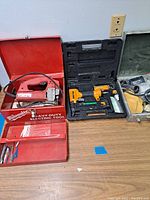 Photo of the three power tools and their cases laid out on a table: a Milwaukee jig saw in red metal case, a black plastic case with yellow Bostitch fastener tool, and a metal case with Porter Cable sander and pads.