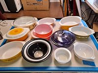 Overhead view showing large assortment of vintage Pyrex casseroles, mixing bowls, in various colors and patterns arranged on a blue edged table. Multiple bowls with and without lids shown.