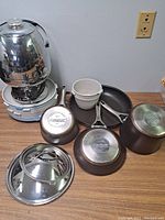 Full view of the kitchen items including the vintage percolator, frying pans, mixing bowls, plastic bowls, and glass lid on a wooden surface.