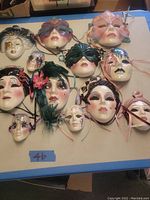 Overhead image of 12 porcelain and ceramic face masks laid out on flat surface with visible ribbons and floral feather embellishments