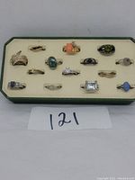 Wide shot of the 14 rings displayed on cream jewelry display board inside green case, shows variety of styles and stones.