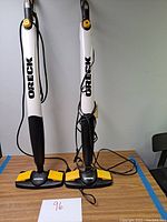 Two Oreck steam floor cleaners standing side by side on a table. Both have white bodies with black bases and yellow control accents. Power cords are visible wrapped around handles.