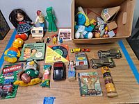 Photo showing a variety of vintage toys including dolls, car coin banks, Fisher Price musical toy, NFL themed plush, Bart Simpson figure, and miscellaneous other items.