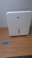 Front view of white Vremi dehumidifier showing logo and water level window with power cord on side.