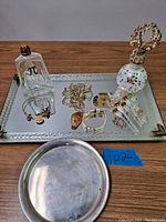 Rectangular glass mirror tray with rope-pattern border holding perfume bottles and jewelry; round sterling silver tray in foreground
