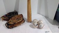 Baseball bat standing vertically alongside two gloves and four baseballs arranged on a surface