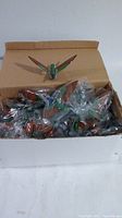 Open cardboard box filled with numerous individually wrapped bird magnets with green bodies, brown wings, and blue heads, one magnet placed outside the packaging on box edge.