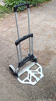 Image showing the folding dolly standing on an asphalt surface, extendable handle upright, base platform flat with metal cutout design.