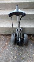 Golf push cart front view showing adjustable handle and wheel assembly on pavement with concrete steps in background.