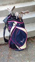 Side angle showing the whole navy blue golf bag with pink and cream accents, standing upright with iron clubs visible inside.