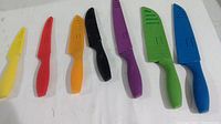 Seven Tomodachi kitchen cutting tools arranged side by side with color-coded plastic handles and matching protective sheaths in yellow, red, orange, black, purple, green, and blue. All tools show normal wear.