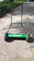 Front view of manual push reel mower showing green blade cylinder, black handle, and wheels with orange rims.